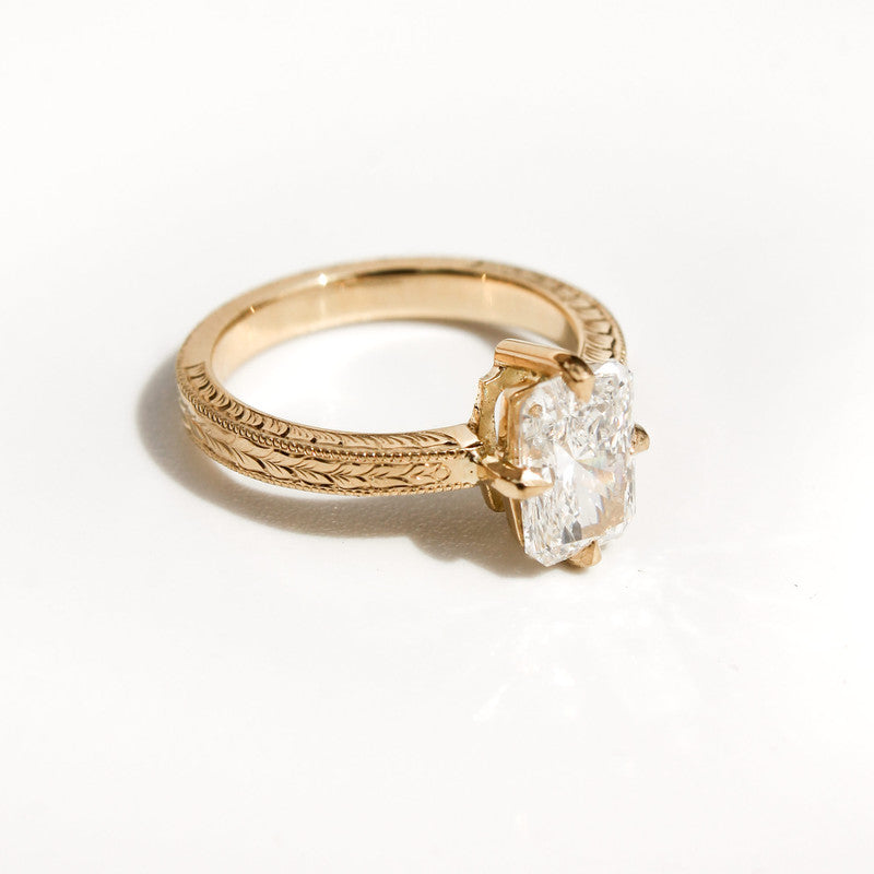 Anillo de oro amarillo con grabados artesanales y diamante rectangular brillante sobre fondo blanco.